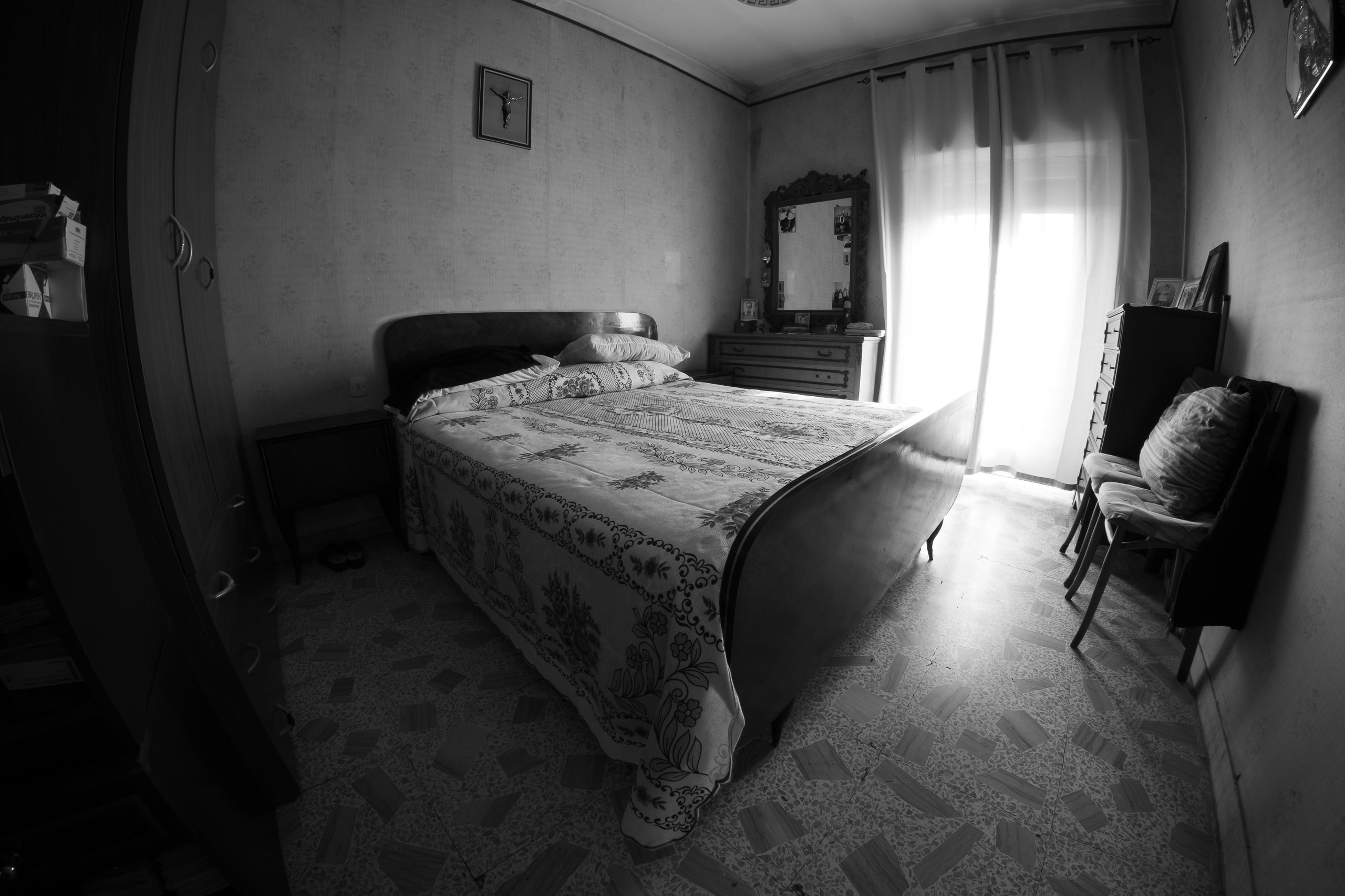 camera da letto con letto matrimoniale, comò con specchio, crocifisso alla parete e luce dalla finestra