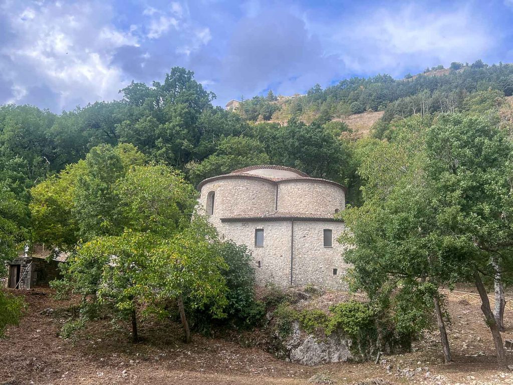retro della chiesa con volumi curvi in pietra e due livelli di copertura, visto da valle tra le querce