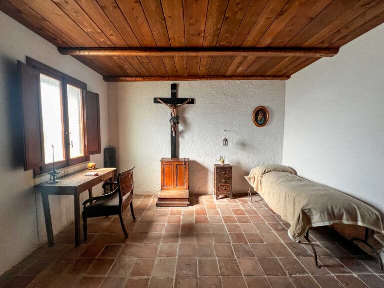 Stanza spoglia con letto singolo, scrittoio con un teschio e inginocchiatoio sotto un grande crocifisso di legno.