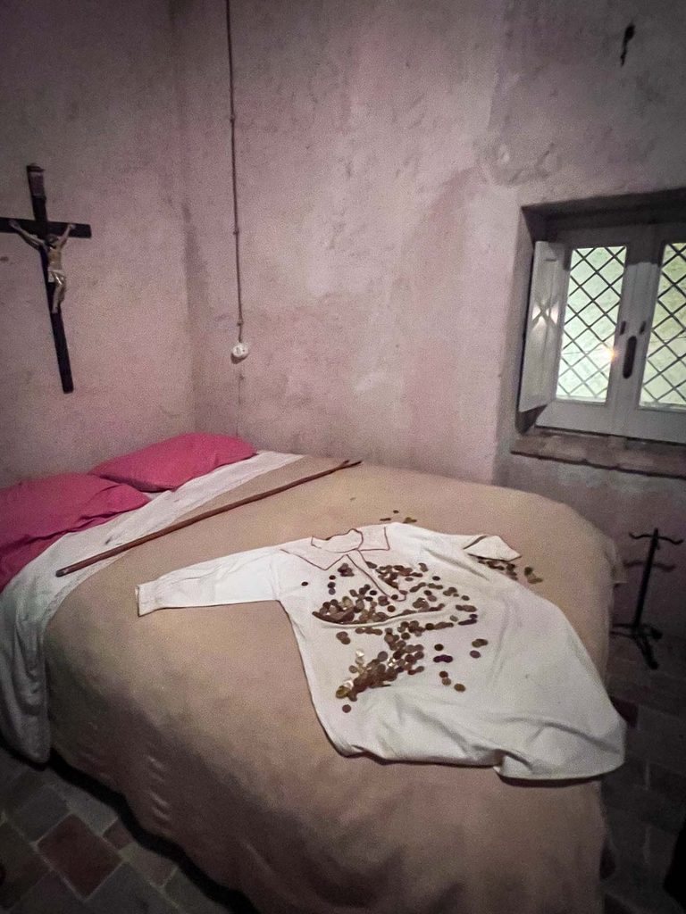 Stanza con letto matrimoniale, crocifisso alla parete, bastone appoggiato al letto e una camicia bianca ricoperta di monete, accanto a una finestra a vetri romboidali.