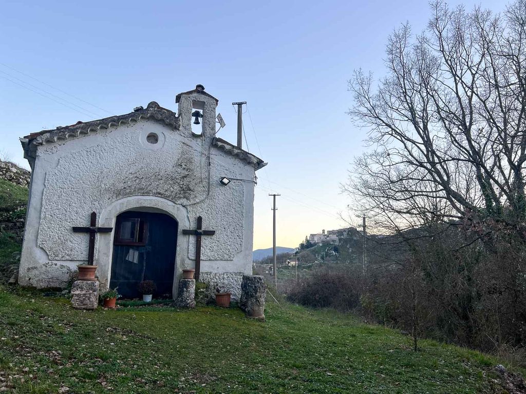 Piccola cappella bianca con campanile a vela e due croci di legno ai lati della porta, con un abitato sulla collina sullo sfondo.
